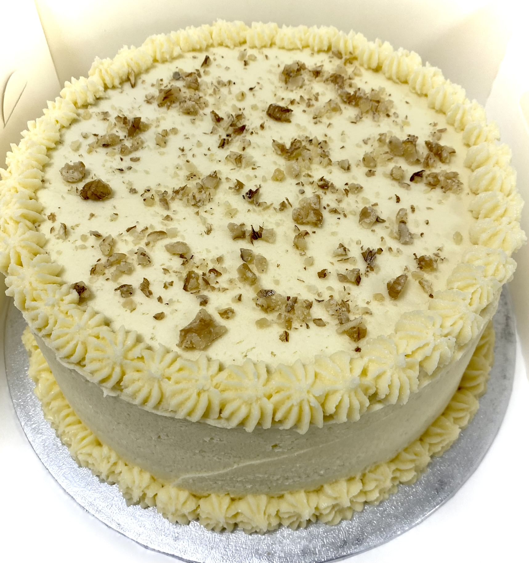A round cake is decorated with white buttercream frosting, featuring piped rosettes around the top edge and the base. The award-winning carrot cake's top surface is sprinkled with chopped nuts, giving it a textured, nutty appearance. This Great Taste Award winner sits on a silver cake board.